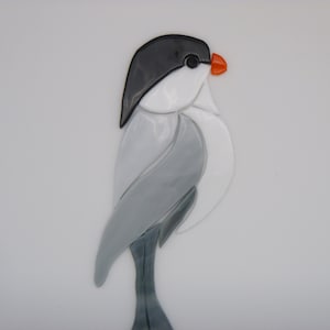 May include: A gray and white glass mosaic bird with a black head and an orange beak. The bird is standing on a gray glass base.