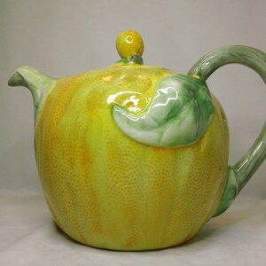May include: A yellow ceramic teapot shaped like a lemon with a green leaf and a yellow knob on the lid.