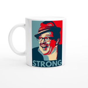 May include: White ceramic mug with a portrait of a man in glasses and a hat, in red, blue, and beige. The word "STRONG" is printed in white at the bottom. A humorous design for a coffee or tea cup.