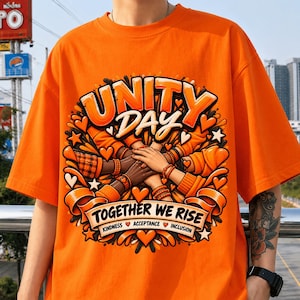 Puede incluir: Una camiseta naranja con un estampado gráfico que incluye el texto UNITY DAY y TOGETHER WE RISE, junto con las palabras KINDNESS, ACCEPTANCE e INCLUSION. El diseño muestra varias manos apiladas, rodeadas de corazones y estrellas naranjas.