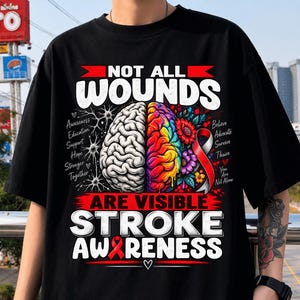 May include: A black t-shirt featuring a graphic of a brain split into a grey side and a colourful side with flowers. Text reads: NOT ALL WOUNDS ARE VISIBLE STROKE AWARENESS, along with words like Awareness, Education, Support, Hope, Believe, Advocate, Survive.