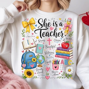 Può includere: Una felpa bianca con una grafica colorata che riporta il testo She is a Teacher, circondata da illustrazioni di un fiocco giallo, una matita, un libro aperto, uno zaino blu, un mappamondo, una pila di libri con una mela e motivi floreali.