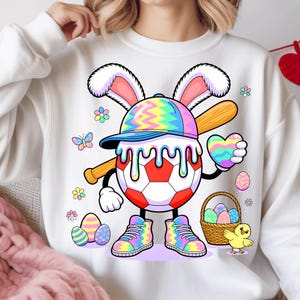 May include: White sweatshirt featuring a cartoon soccer ball character with bunny ears, a baseball bat, and a heart. The character wears a baseball cap and sneakers with rainbow patterns. Easter eggs, flowers, and a chick are also shown.