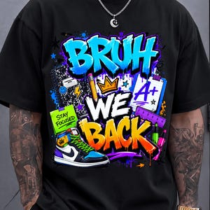 May include: A black t-shirt featuring a colorful graphic print with the text "BRUH WE BACK". The design includes a blue graffiti-style font, a yellow crown, a notebook, a green sticky note with "STAY FOCUSED", a blue paper with "A+", and a sneaker.