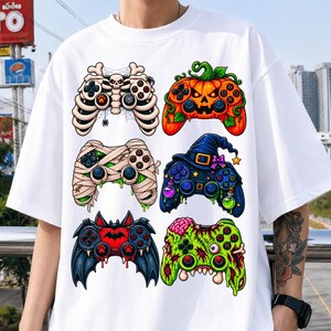 Op de afbeelding: Een wit t-shirt met een grafische print van zes videogamecontrollers in Halloween-thema. De ontwerpen bevatten een skelet, een pompoen, een mummie, een heks met een hoed, een vleermuis en een groene zombie met zichtbare hersenen.