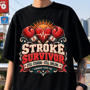 May include: A black t-shirt featuring a graphic design with two red boxing gloves, a central heart with an EKG line, and a sunburst. The text reads STROKE SURVIVOR, STILL STANDING STILL SMILING, and STRONGER EVERY DAY with red ribbons.