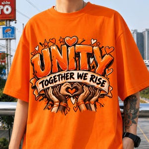 Puede incluir: Una camiseta de manga corta de color naranja brillante con un estampado gráfico que incluye el texto "UNITY TOGETHER WE RISE" en letras naranjas llamativas. El diseño muestra varias manos de distintos tonos de piel formando un corazón.