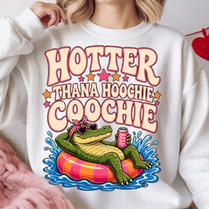 May include: White sweatshirt featuring the text "HOTTER THANA HOOCHIE COOCHIE" in pink and orange. A cartoon alligator wearing sunglasses and a pink bandana is relaxing on an inflatable ring in water. The alligator holds a can with the words "Lake Hair Don't Care."