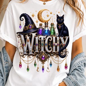 May include: A white t-shirt featuring a graphic print of the word WITCHY in ornate silver lettering. The design includes a black witch hat, a black cat, a golden crescent moon, stars, and colorful potion bottles, all adorned with hanging crystals.