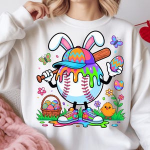 Puede incluir: Sudadera blanca con un diseño de béisbol de Pascua colorido. La pelota de béisbol tiene orejas de conejo, un bate y sostiene un huevo decorado. El diseño incluye una gorra de béisbol, gotas coloridas, mariposas, flores, un pollito y una cesta de huevos.