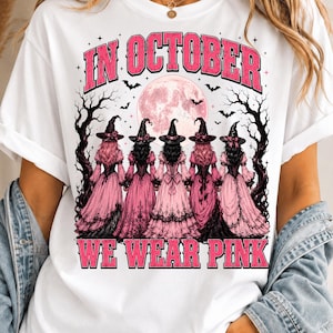 以下が含まれることがあります： 白いTシャツに、ピンクのドレスと黒い帽子を身につけた5人の魔女が、満月と枯れ木を背景に立っているグラフィックが描かれています。文字はピンクの太字で「IN OCTOBER WE WEAR PINK」と書かれています。デザインには小さな黒いコウモリと星が含まれています。