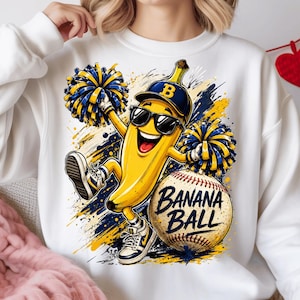 May include: White sweatshirt featuring a cartoon banana character wearing sunglasses and a baseball cap, holding pom-poms, and sneakers. A baseball with the text "BANANA BALL" is next to the banana. The design includes blue and yellow accents.