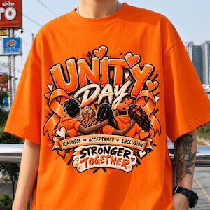 Puede incluir: Una camiseta de color naranja brillante con un estampado gráfico que incluye el texto UNITY DAY, KINDNESS, ACCEPTANCE, INCLUSION y STRONGER TOGETHER. El diseño muestra a cinco personas abrazadas, rodeadas de cintas naranjas y corazones.