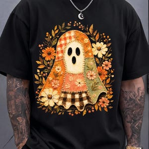 Può includere: Una t-shirt nera con una grafica centrale raffigurante un fantasma realizzato con motivi in tessuto patchwork, inclusi quadretti vichy arancioni e stampe floreali. Il fantasma è circondato da una corona di fiori e foglie dai toni autunnali.
