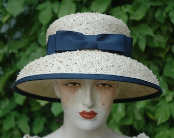 Puede incluir: Un sombrero de paja blanco con una cinta y un ribete azul marino. El sombrero tiene una ala ancha y está decorado con pequeñas cuentas blancas.