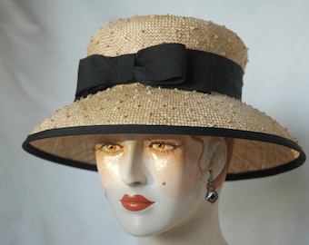 Puede incluir: Un sombrero de paja de ala ancha con una cinta negra atada en un lazo. El sombrero está decorado con pequeñas cuentas espaciadas uniformemente.