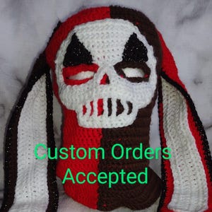 Peut inclure: Un masque de crâne au crochet fait à la main en rouge, blanc et marron. Le masque présente un motif de crâne avec des accents noirs et de longues oreilles tombantes. Le texte "Custom Orders Accepted" est visible.