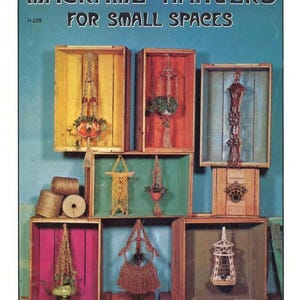 May include: A vintage instructional booklet cover titled MACRAME' HANGERS FOR SMALL SPACES, featuring various macrame plant hangers displayed inside colorful wooden crates. The image includes spools of twine and the text H-228 and 30-13117.
