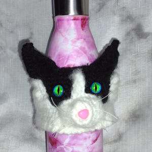 May include: A pink and white marble-effect water bottle with a stainless steel lid. It has a handmade cat cosy with black ears, a white face, green eyes, and a pink nose. The cosy adds a playful touch to the bottle.