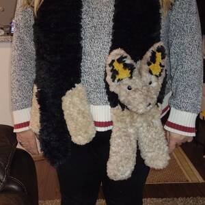 May include: A grey and white striped sweater with a black and tan scarf featuring a plush animal design. The sweater has white cuffs with red stripes. The scarf has a plush animal head with yellow accents.
