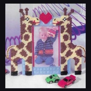 May include: A plastic canvas picture frame featuring two brown and cream giraffes on either side of a central rectangular opening. A red heart is positioned at the top center. The frame holds a photograph of a child wearing a patterned sweater.