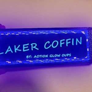 May include: A rectangular, black device with the text "LAKER COFFIN" in light blue, illuminated by a blue glow. The text "BY: ACTION GLOW CUPS" is below. A switch and wires are visible on the side.