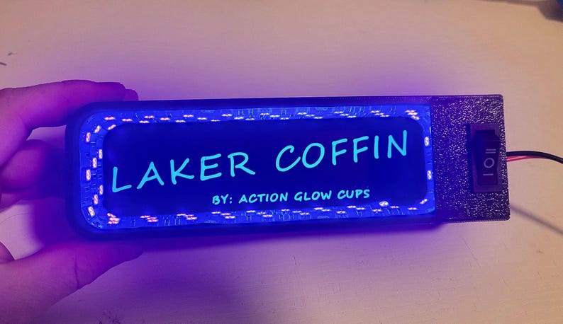 May include: A rectangular, black device with the text "LAKER COFFIN" in light blue, illuminated by a blue glow. The text "BY: ACTION GLOW CUPS" is below. A switch and wires are visible on the side.