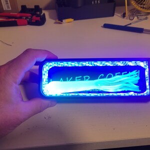 May include: A rectangular device with a blue LED light frame, illuminating a blue interior with the text "LAKER COFFEE" and a bundle of light blue fibers. A switch is on the right side.
