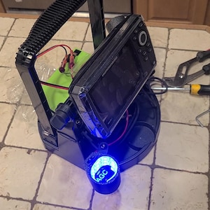 May include: A black electronic device with a screen, mounted on a black base with a blue light. A green battery pack and red wires are visible. Pliers, a screwdriver, and an Allen wrench are nearby.