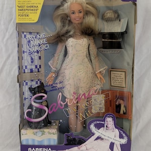 May include: A Sabrina the Teenage Witch doll in original packaging, featuring a blonde doll in a white dress, a black cat figure, a top hat, and a book. The box includes text such as Try Me... Make Magic!, Salem the Cat with Glowing Eyes, and Sabrina The Teenage Witch, with a photo of the doll and a sweepstakes promotion.