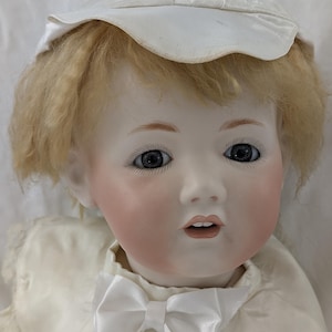May include: A vintage porcelain doll with blonde hair and blue eyes, wearing a white satin outfit and a matching cap. The doll has rosy cheeks and a white bow tie. The doll is a collectible item.