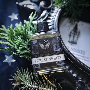 May include: A small glass bottle with a black label that reads "House of Moth & Stars" and "Forest Nights". The label features a black and white illustration of a moth with a crescent moon and stars. The bottle is sitting on a dark blue surface with green foliage.