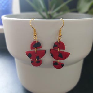 Könnte beinhalten: Ein Paar abstrakte rote und schwarze Ohrringe mit goldfarbenen Haken. Die Ohrringe bestehen aus rotem Material mit schwarzen Tupfen. Die Ohrringe hängen vor einem weißen Topf mit einer grünen Pflanze.