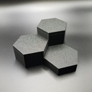 May include: Three black, hexagonal display boxes with textured, gray tops are stacked in a pyramid formation. The boxes are made of a matte material and sit on a dark surface, creating a modern, minimalist aesthetic.