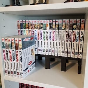 Può includere: Una collezione di volumi manga Claymore di Norihiro Yagi, disposti su uno scaffale bianco. Il set include i volumi dal 7 al 21 disposti verticalmente in fila, con i volumi dal 22 al 27 esposti frontalmente, mostrando il testo e le illustrazioni.