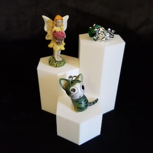 May include: A display of small figurines and jewelry on white geometric pedestals. A yellow-clad fairy holds a basket of red flowers. A silver ring with green stones and a green and white cat figurine are also displayed.