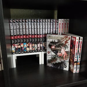 Può includere: Una collezione di libri manga disposti su uno scaffale. Il set include i volumi da 0 a 16 di Jujutsu Kaisen con dorsi neri e i volumi 5, 6 e 7 di Sakamoto Days. Il volume 5 di Sakamoto Days è esposto frontalmente con un'illustrazione.