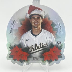 Könnte beinhalten: Baseballkarte mit Jacob Wilson von den Athletics, mit Weihnachtsmütze. Die Karte hat ein Poinsettien- und Grünpflanzen-Design. Das Athletics-Logo und die Marke Topps sind sichtbar.
