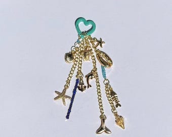 Könnte beinhalten: Ein türkisfarbener herzförmiger Anhänger mit einer goldenen Kette und verschiedenen goldenen Anhängern, darunter eine Muschel, ein Delfin, ein Seestern, ein Anker, ein Fisch und ein Meerjungfrauen-Schwanz.