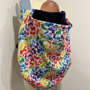 May include: A baby carrier with a colorful butterfly pattern. The carrier features a white background with a vibrant array of butterflies in shades of red, yellow, blue, green, and purple. The carrier has a blue denim strap and a dark blue hood.