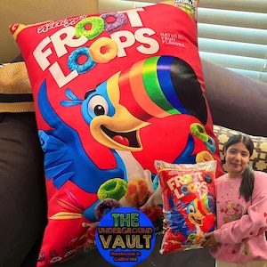 Könnte beinhalten: Ein großes rotes Kissen mit einem Tukan und dem Aufdruck "Froot Loops". Das Kissen wird von The Underground Vault hergestellt und ist handgefertigt in Kalifornien. Eine Person hält eine Tüte Froot Loops Müsli.