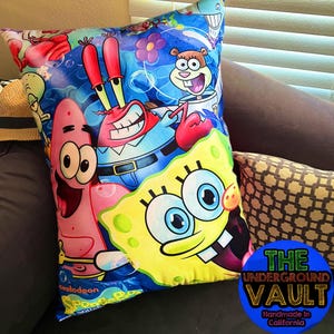 Puede incluir: Una almohada colorida con personajes de la caricatura Bob Esponja. La almohada es azul con acentos amarillos, rosados, rojos y marrones. Los personajes incluyen Bob Esponja, Patricio Estrella, Calamardo Tentáculos y Don Cangrejo. La almohada tiene el logotipo de Nickelodeon y las palabras "Bob Esponja" impresas en ella.