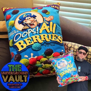 Puede incluir: Una almohada azul y amarilla con las palabras "Oops! All Berries" y un personaje de pirata de dibujos animados. La almohada está hecha por The Underground Vault y está hecha a mano en California.