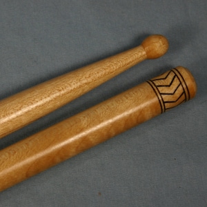 May include: Two wooden drumsticks, one with a plain round tip and the other with a decorative black chevron pattern near the tip.