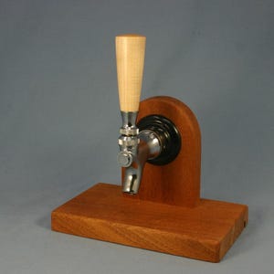 May include: A wooden beer tap with a light-colored handle and a stainless steel tap. The tap is mounted on a rectangular wooden base and a curved wooden support. The wood has a reddish-brown hue.