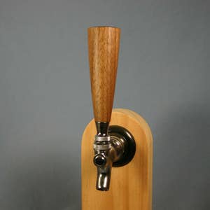 May include: A wooden beer tap handle with a conical shape, mounted on a light-colored wooden base. The tap features a polished metal spout and a dark metal collar, set against a gray background.