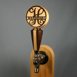 May include: Wooden tap handle with a circular top featuring the name "HARDESKI" and a decorative initial. The handle has a dark wood and light wood design, with a metal tap. The tap is mounted on a wooden base.