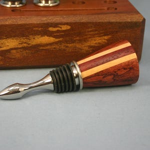May include: A wine bottle stopper featuring a conical handle made of alternating dark reddish-brown and light tan wood segments. The stopper has a polished silver-tone metal tip and a black ribbed silicone seal for a secure fit in a bottle.