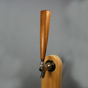 May include: A beer tap handle featuring a tall, tapered, medium-brown wooden grip attached to a silver-colored metal faucet mechanism. The faucet is mounted onto a light-colored, rounded wooden base, creating a functional beverage dispensing unit.