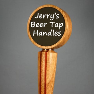 May include: A wooden beer tap handle with a round top and a rectangular base. The top has a black circular insert with the text "Jerry's Beer Tap Handles" in white. The handle is made of light and dark wood.